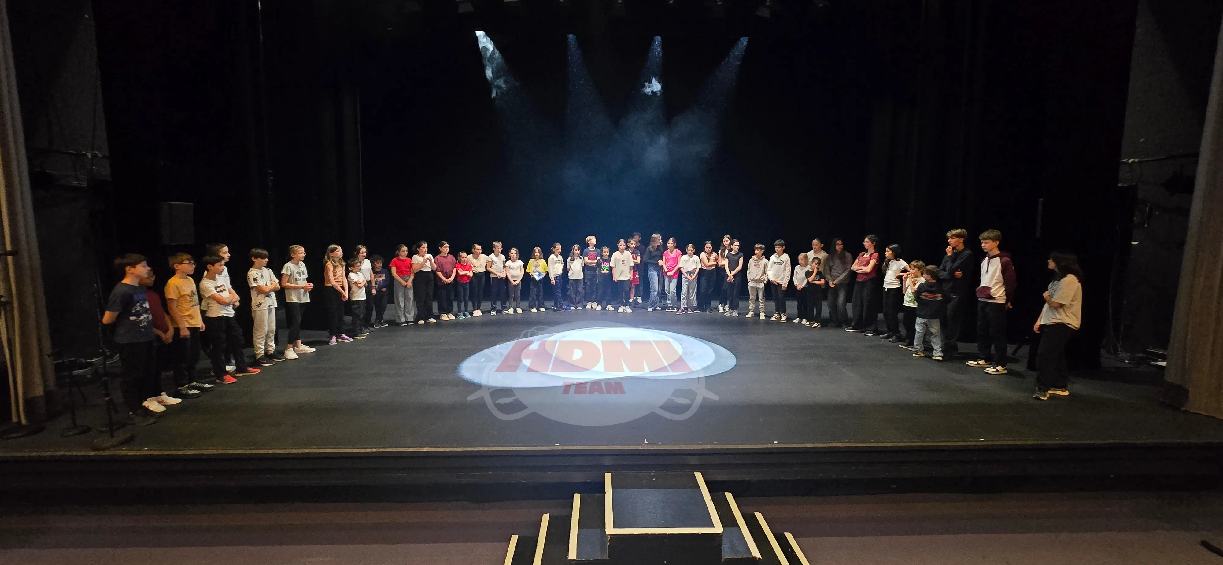 Groupe de jeunes réuni en cercle sur une scène de théâtre à l'issue d'une intervention HDMI Team, logo HDMI TEAM projeté au sol