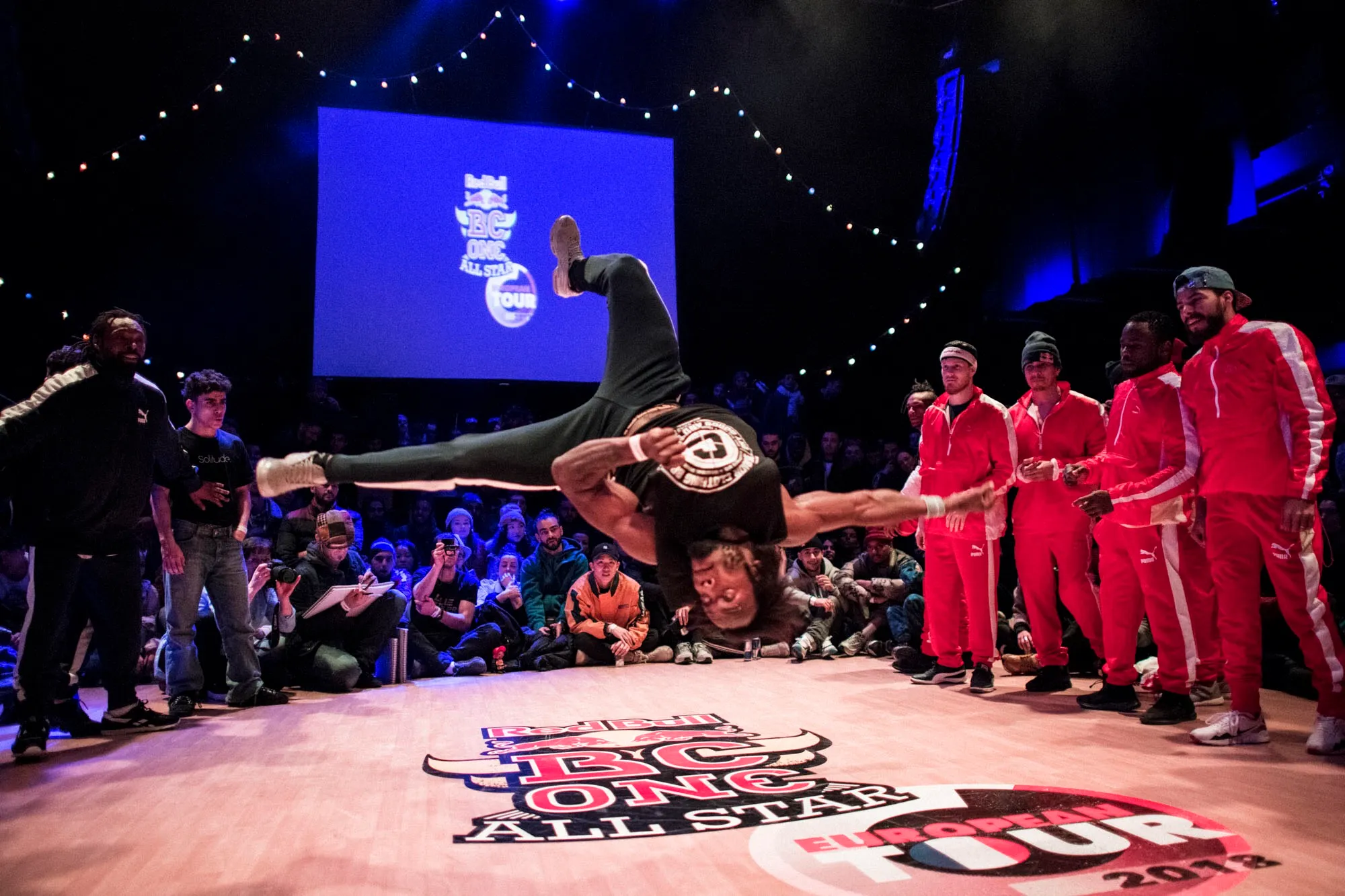 Julee-One en figure aérienne lors d'un battle Red Bull BC One All Star European Tour, entouré du public et des autres danseurs